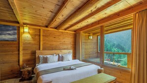 A wooden cabin bedroom with a bed, bedside table, bench, and large windows offering a forest view. - ELIVAAS Silvana 3 BHK Cottage  (Kumharsain)