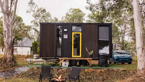 A small, dark-colored mobile home with a yellow door, a fire pit, and two chairs in the foreground.