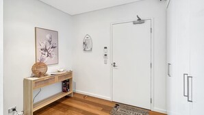 A modern room with a wooden console table, a framed floral artwork, a white door with a peephole, and a welcome mat. - Stylish Apartment Near Mandurah’s Best Spots (Mandurah)
