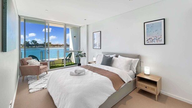 A modern bedroom with a large bed, a small bedside table, a chair, and a view of the sea through a sliding glass door. - Stylish Apartment Near Mandurah’s Best Spots (Mandurah)