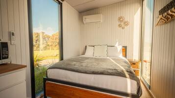 A bedroom with a bed, a bedside table, an air conditioning unit, and a view of the outdoors.