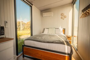 A bedroom with a bed, a bedside table, an air conditioning unit, and a view of the outdoors. - Rimu View 4 by Tiny Away (Ruatapu)