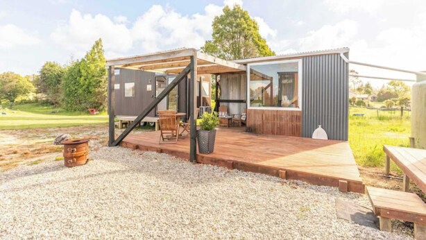 A modern cabin with a wooden deck, outdoor seating, and a large window overlooking a grassy area.