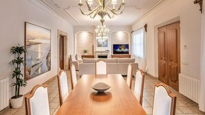 Una sala da pranzo con un tavolo in legno, sedie bianche, un lampadario, una televisione e un divano.