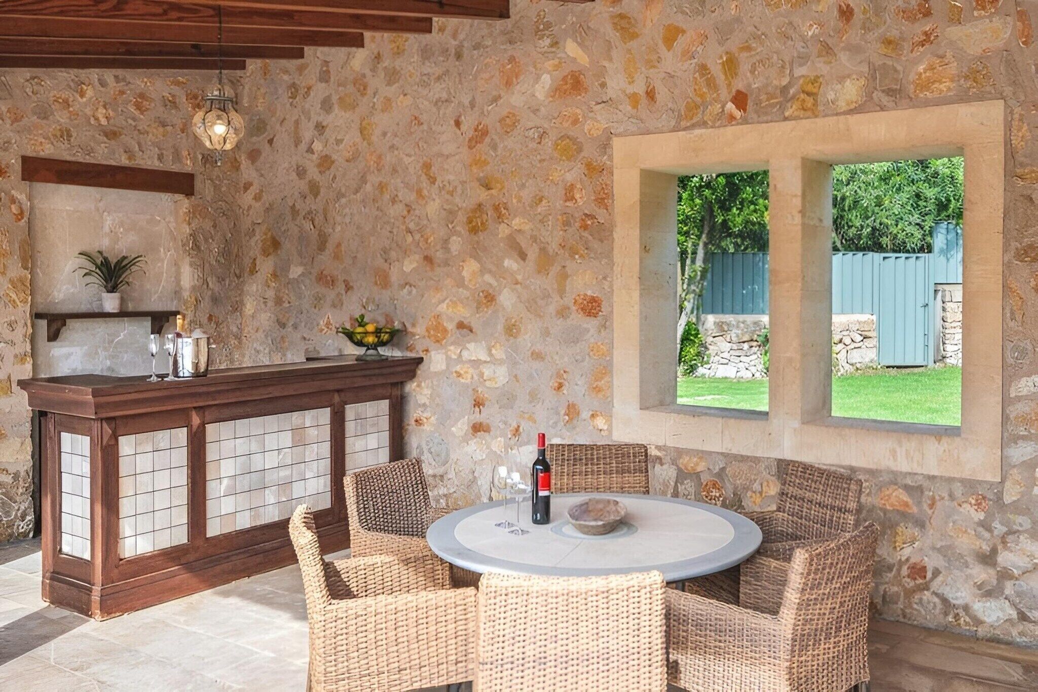 Un espace extérieur aménagé pour les repas, avec des murs en pierre, des chaises en osier et une table ronde, un bar doté d’un comptoir en pierre, et une vue sur une pelouse verdoyante par le biais d’une fenêtre.