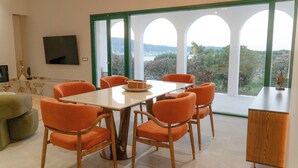 Ein Essbereich mit Tisch und Stühlen, einem Fernseher an der Wand und großen Fenstern mit Blick ins Grüne.