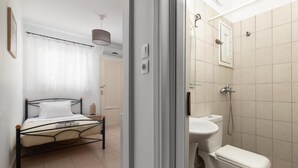 A bedroom with a bed, a bedside table, a hanging lamp, and a bathroom with a toilet and shower. - Amethyst Rooms (Rhodes)