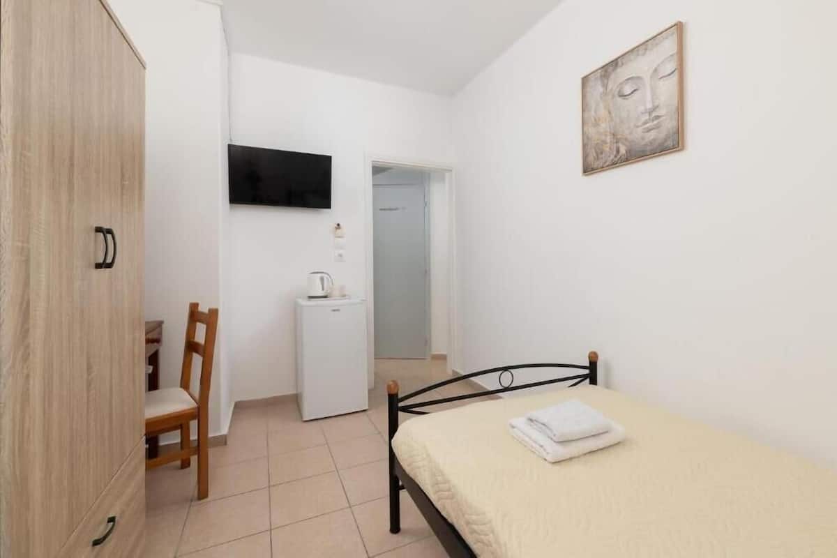 A bedroom with a bed, a wooden wardrobe, a desk, a chair, and a television on the wall.
