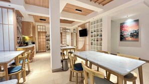 A dining area with wooden tables and chairs, a TV mounted on the wall, and a bookshelf filled with books. - Jambuluwuk Mansion Adityawarman (Jakarta)