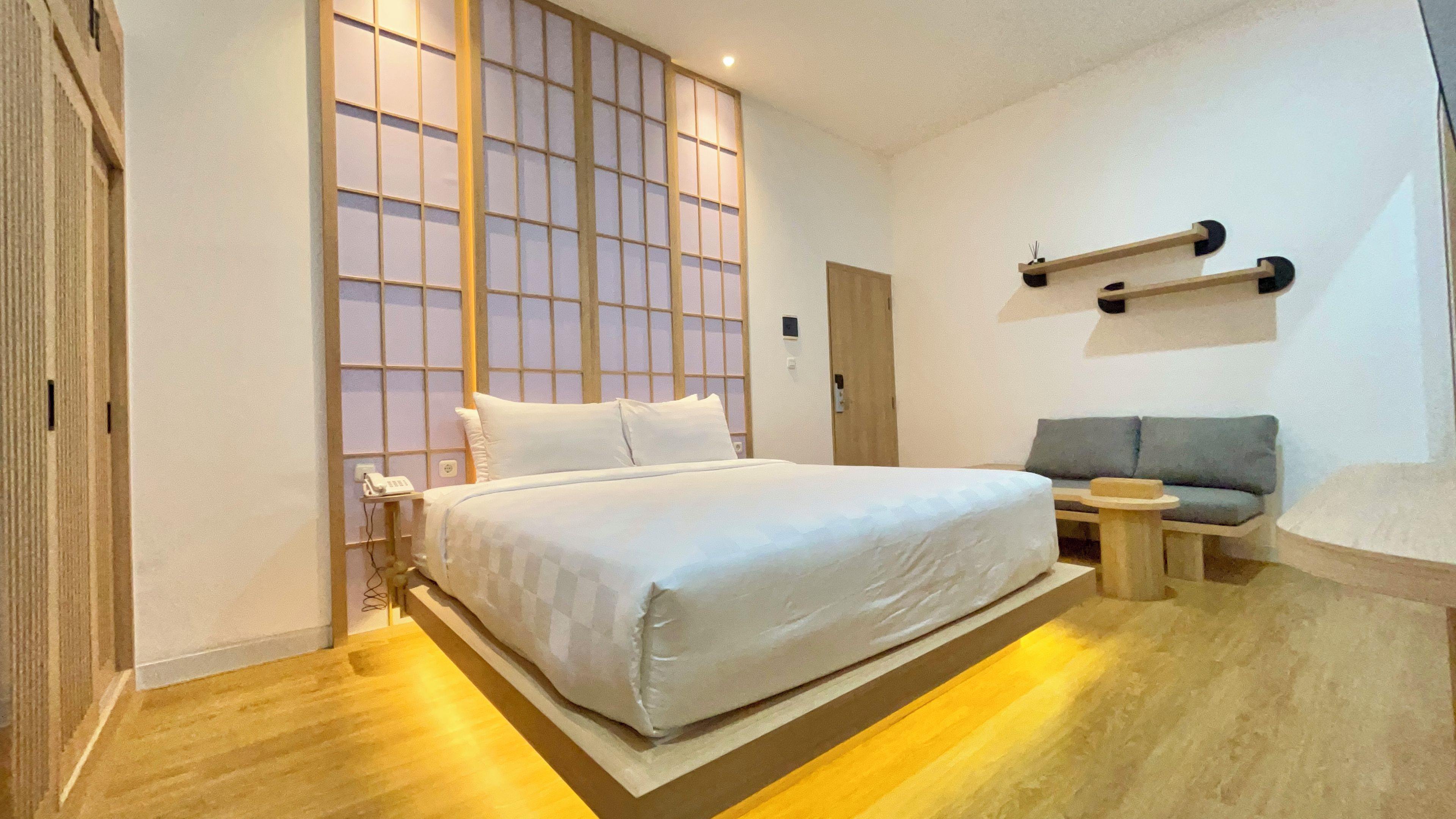 A hotel room with a bed, a small seating area, a wall-mounted shelf, and a sliding door with a view.