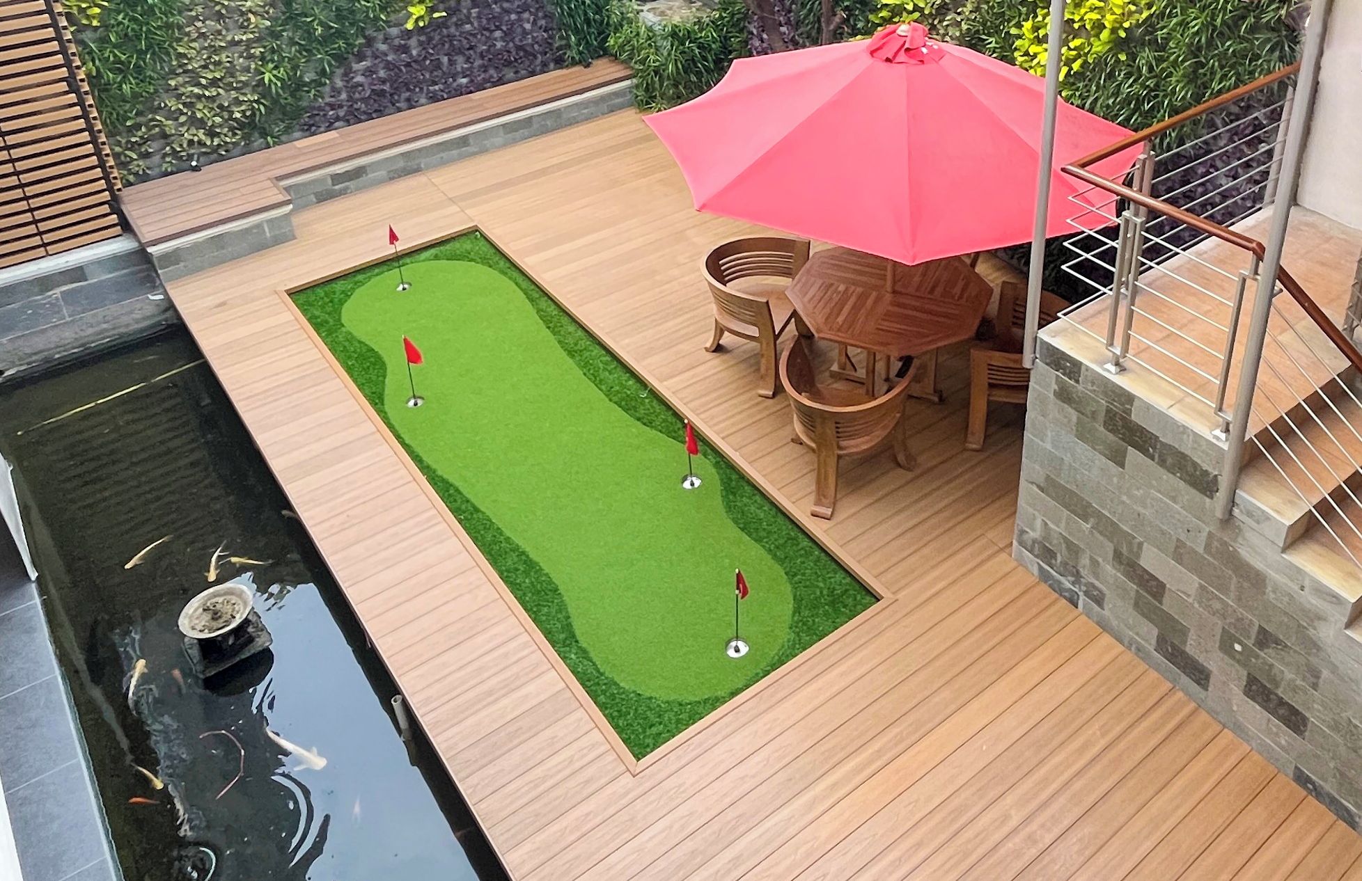 Eine Holzterrasse mit Putting Green, einem kleinen Teich und einem roten Sonnenschirm.