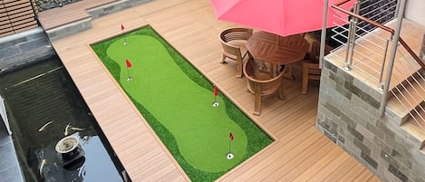 A wooden deck with a putting green, a small pond, and a red umbrella.