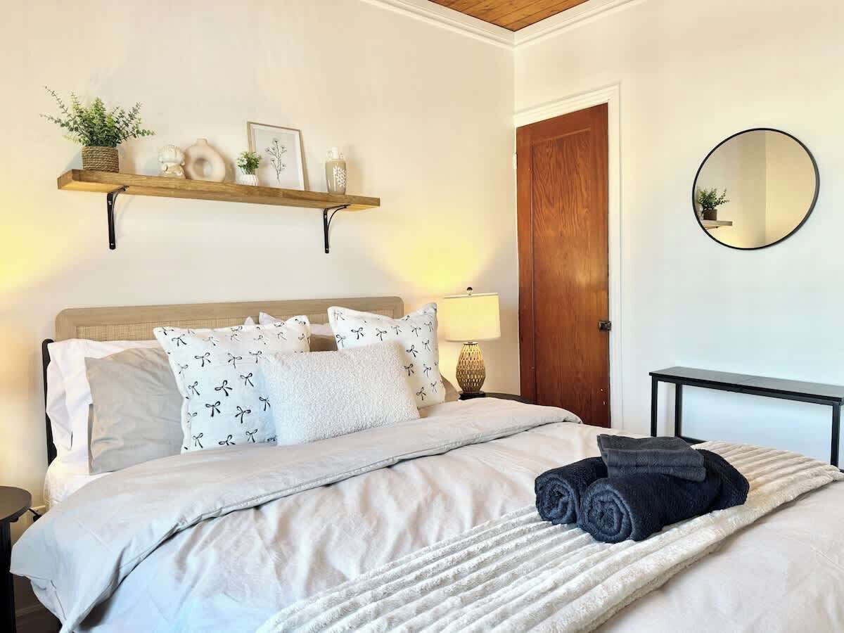 Cama bem arrumada com esquema de cores branco e cinza, cabeceira de madeira, mesa de cabeceira com abajur e prateleira na parede com objetos decorativos.