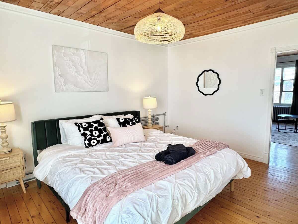 A bedroom with a wooden ceiling, a bed with white bedding and a pink throw, a bedside lamp, and a wall-mounted mirror.