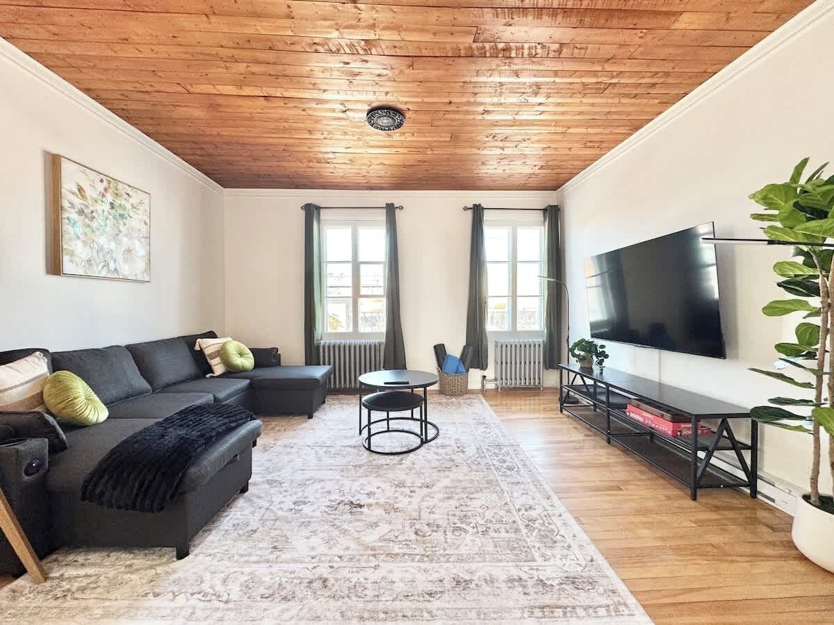 A modern living room with a wooden ceiling, a sectional sofa, a round coffee table, a flat-screen TV, and a large potted plant.
