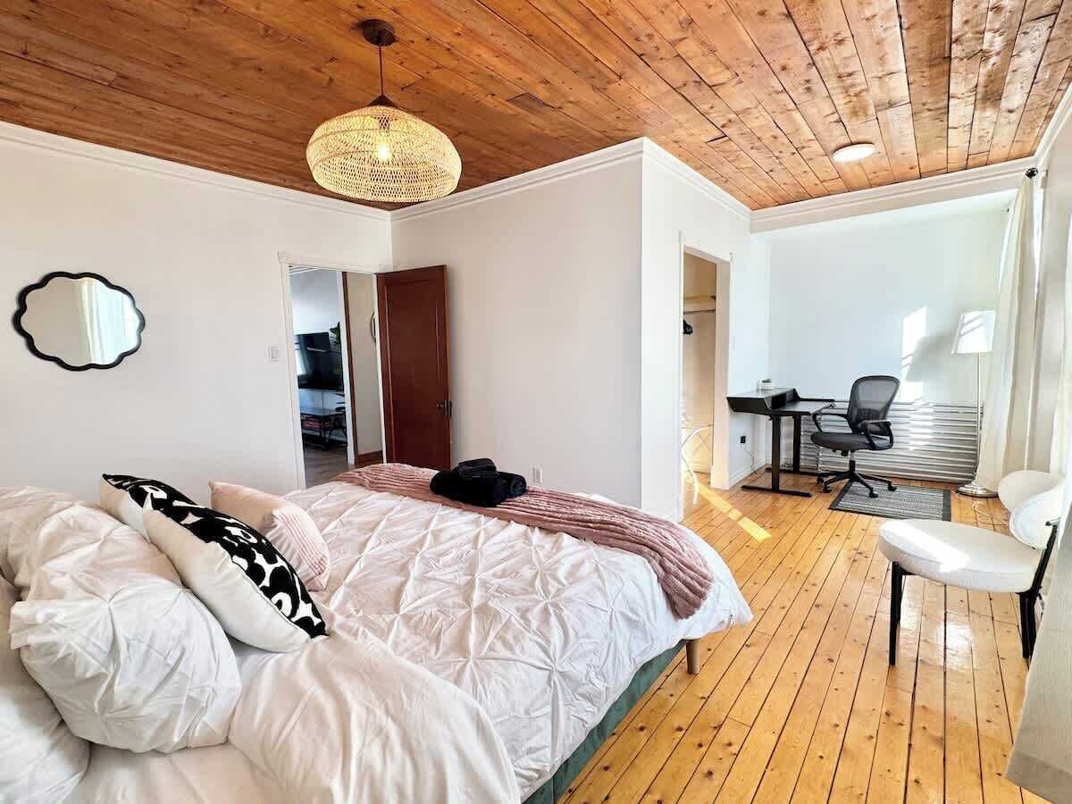 A bedroom with a wooden ceiling, a bed with white bedding, a woven light fixture, a chair, and a desk with a chair.
