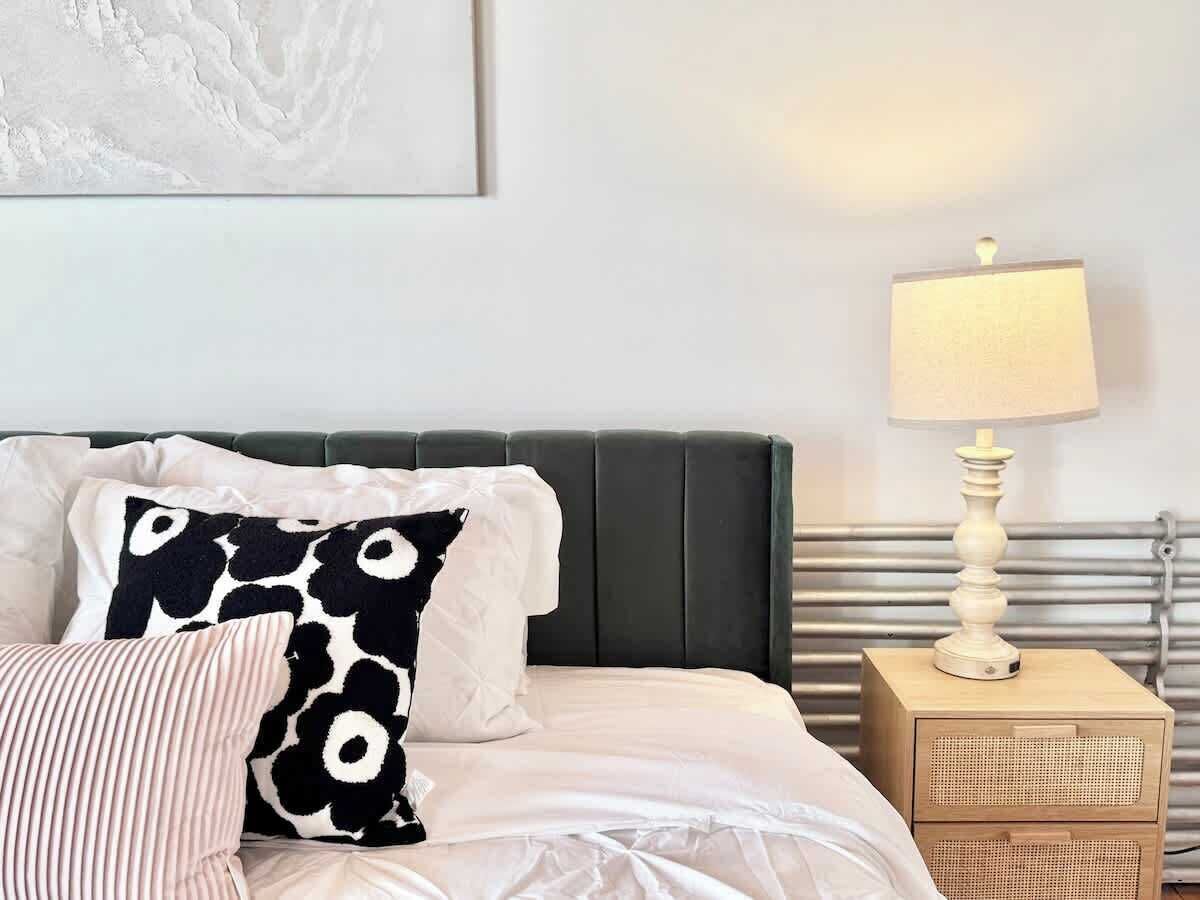 Un lit bien fait, avec une literie blanche, un coussin à motifs noir et blanc et un coussin rayé. On y trouve une table de chevet avec une lampe et une petite enceinte.