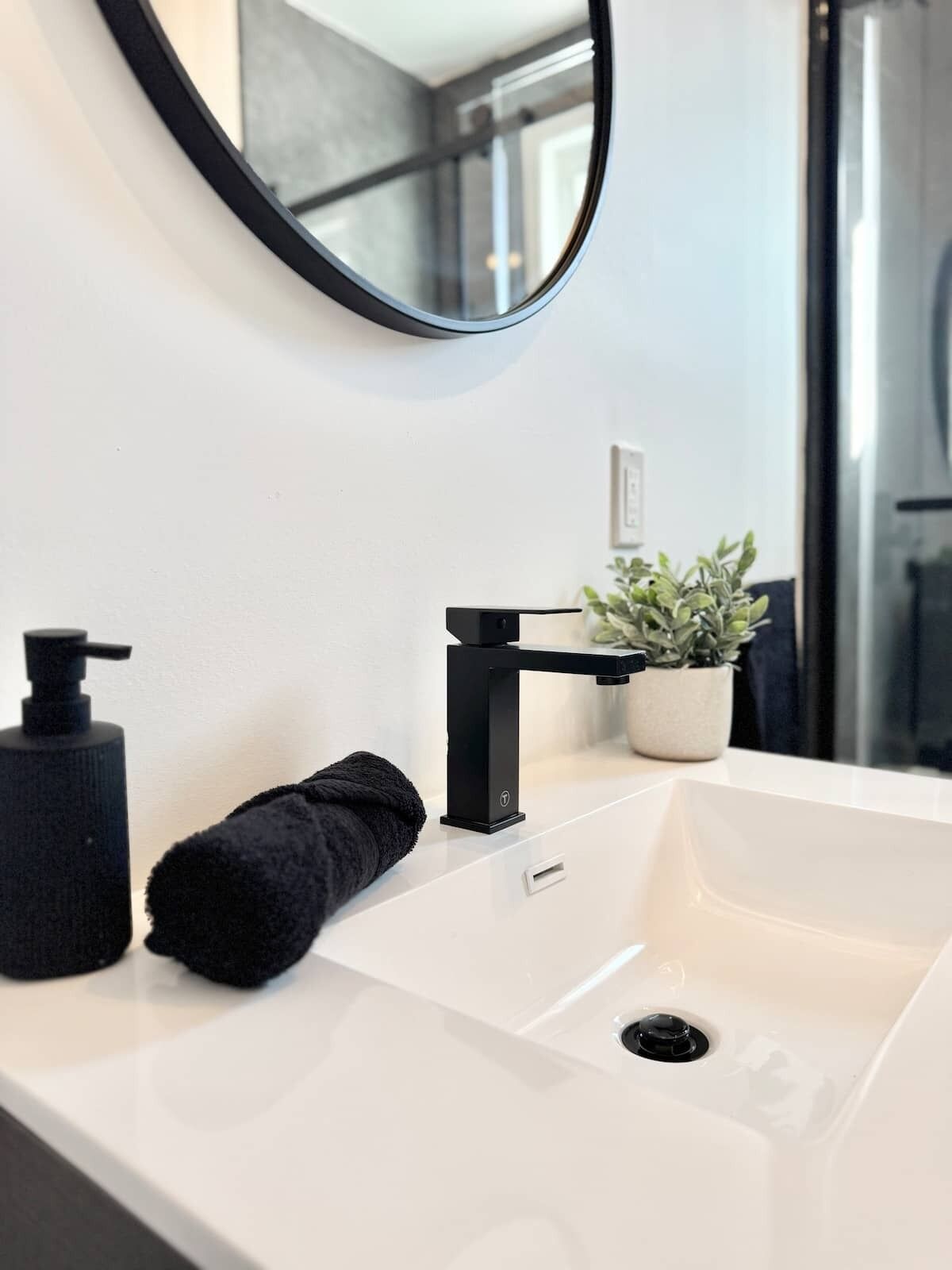 Un bagno con un lavandino bianco, un rubinetto nero e uno specchio rotondo. Sul piano di appoggio c'è un asciugamano nero e una pianta in vaso.