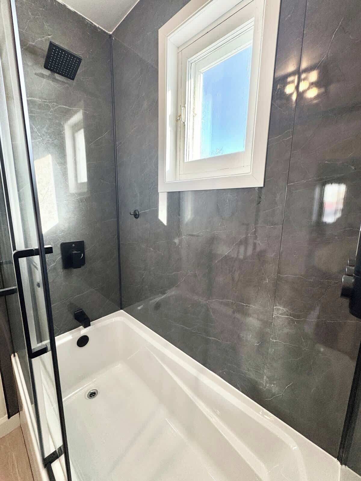 A modern bathroom with a glass-enclosed shower, a white bathtub, and a window providing natural light.