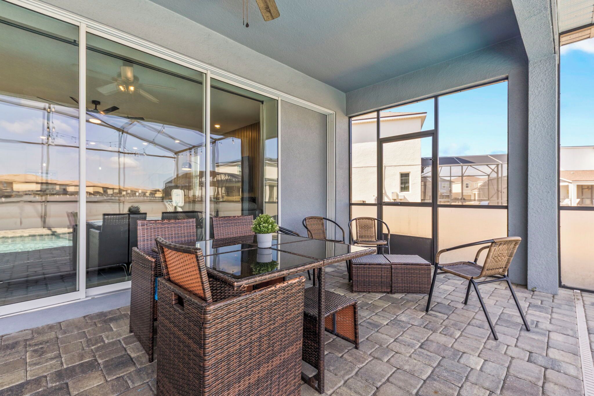 A covered patio area with a glass table and wicker chairs, overlooking a swimming pool and neighboring houses.