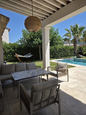 Eine Terrasse im Freien mit einer Couch, Stühlen, einem Tisch, einer Hängelampe und einem Pool.