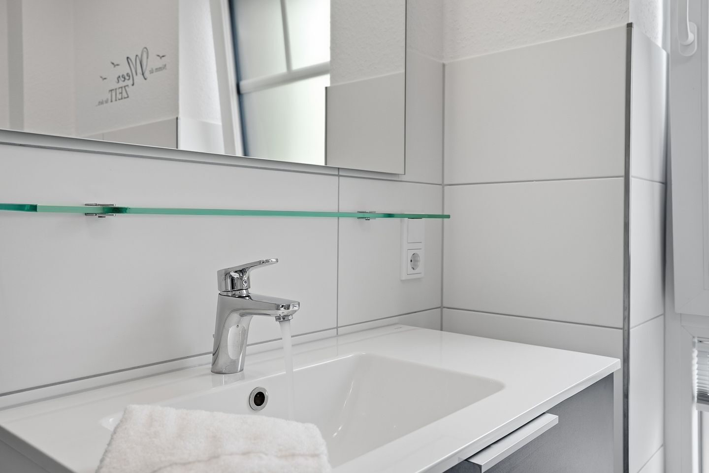 Un baño moderno con un lavamanos blanco, grifo cromado y una repisa de vidrio arriba.