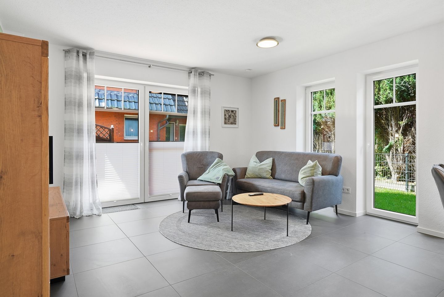 Ein modernes Wohnzimmer mit einem grauen Sofa, einem kleinen runden Tisch und einem grauen Sessel. Eine Glastür öffnet sich zum Balkon mit Blick auf ein Gebäude und Grünflächen.
