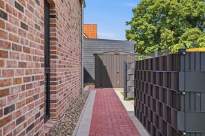 A narrow brick alleyway with a red brick path, a metal fence, and a grey building with a door labeled 'Cordula.' - Cordula - Modern vacation apartment Cordula with garden, terrace and e-charging station (Krummhörn)