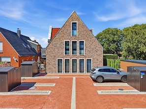 A modern two-story house with a brick facade, a car parked in the driveway, and a clear blue sky. - Cordula - Modern vacation apartment Cordula with garden, terrace and e-charging station (Krummhörn)