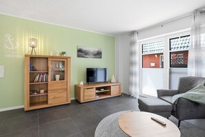 A living room with a television, a bookshelf, a grey sofa, and a round coffee table. - Cordula - Modern vacation apartment Cordula with garden, terrace and e-charging station (Krummhörn)