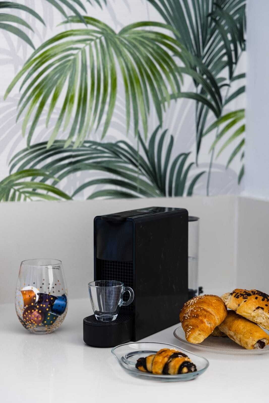 Un comptoir de cuisine avec une machine à café, un verre d’eau, une assiette de croissants et un récipient contenant des objets colorés.
