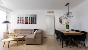 A modern living room with a beige sofa, a round coffee table, and a dining area with a wooden table and black chairs. There is a colorful abstract painting on the wall and a vase with flowers on the dining table. - MAMAD - Bright Eilat escape with balcony n pool (Eilat)