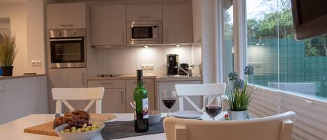 A modern kitchen with white cabinetry, a dining table with chairs, a bottle of wine, and a bowl of fruit.