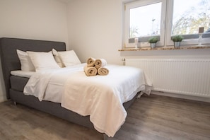 A neatly made bed with rolled towels, a grey sofa, a white radiator, and a window with plants on the sill. - Haus Seestern - Holiday home Seestern with modern furnishings and fenced garden (Cuxhaven)