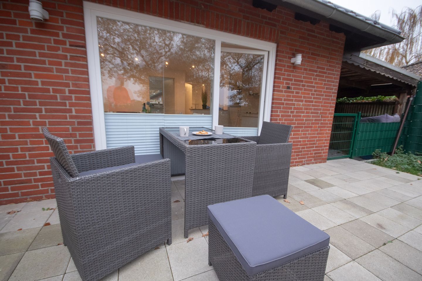 An outdoor patio with wicker furniture and a table, a large window, and a brick wall.