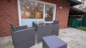 An outdoor patio with wicker furniture and a table, a large window, and a brick wall.