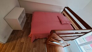 A bedroom with a wooden bed frame, a red bedspread, a wooden nightstand, and a white lamp. - Mezzanine studio full (Bordeaux)