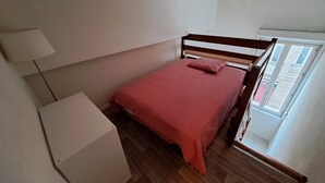 A small, neatly arranged bedroom with a wooden bed, a white bedside table, and a window providing a view of the outside. - Mezzanine studio full (Bordeaux)