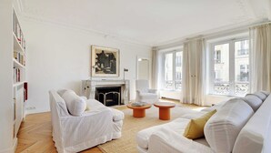 Een ruime woonkamer met een open haard, witte banken en een groot raam met uitzicht op gebouwen.
