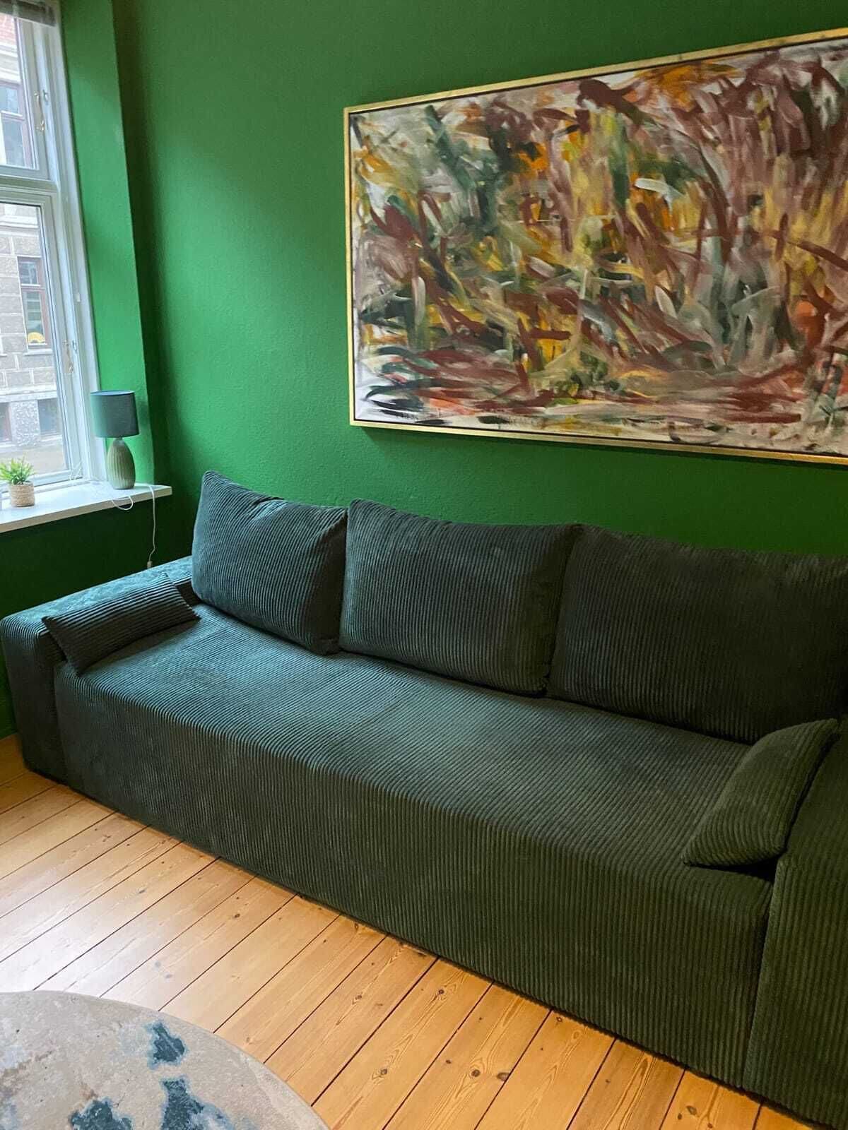 A room with a green wall, a large abstract painting, a dark green sofa with cushions, a small table with a lamp, and a window with a view of buildings.