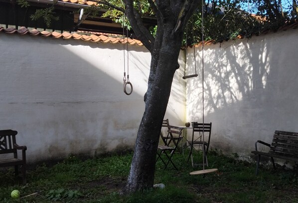 Una zona patio con un muro bianco, un albero e un'amaca. Sono presenti due panchine in legno e un tavolo con sedie.