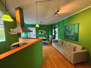 A modern kitchen with a wooden countertop and a built-in stove, a green wall with abstract art, a white sofa, and a wooden floor.