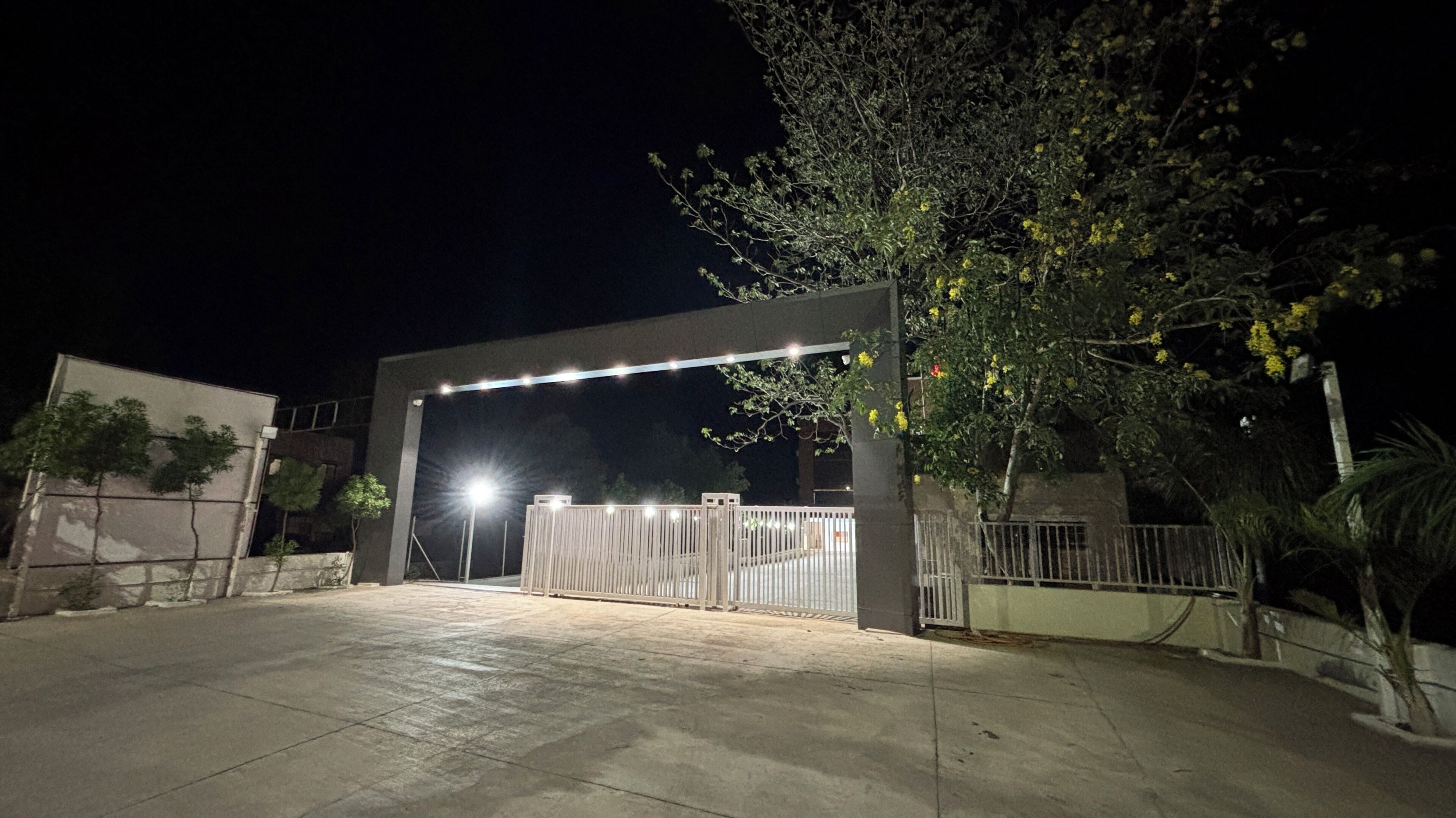 A gated entrance with a metal fence, a tree with yellow flowers, and a building in the background.