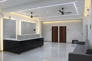 A modern lobby with a reception desk, two wooden doors, a ceiling fan, and a grey sofa.