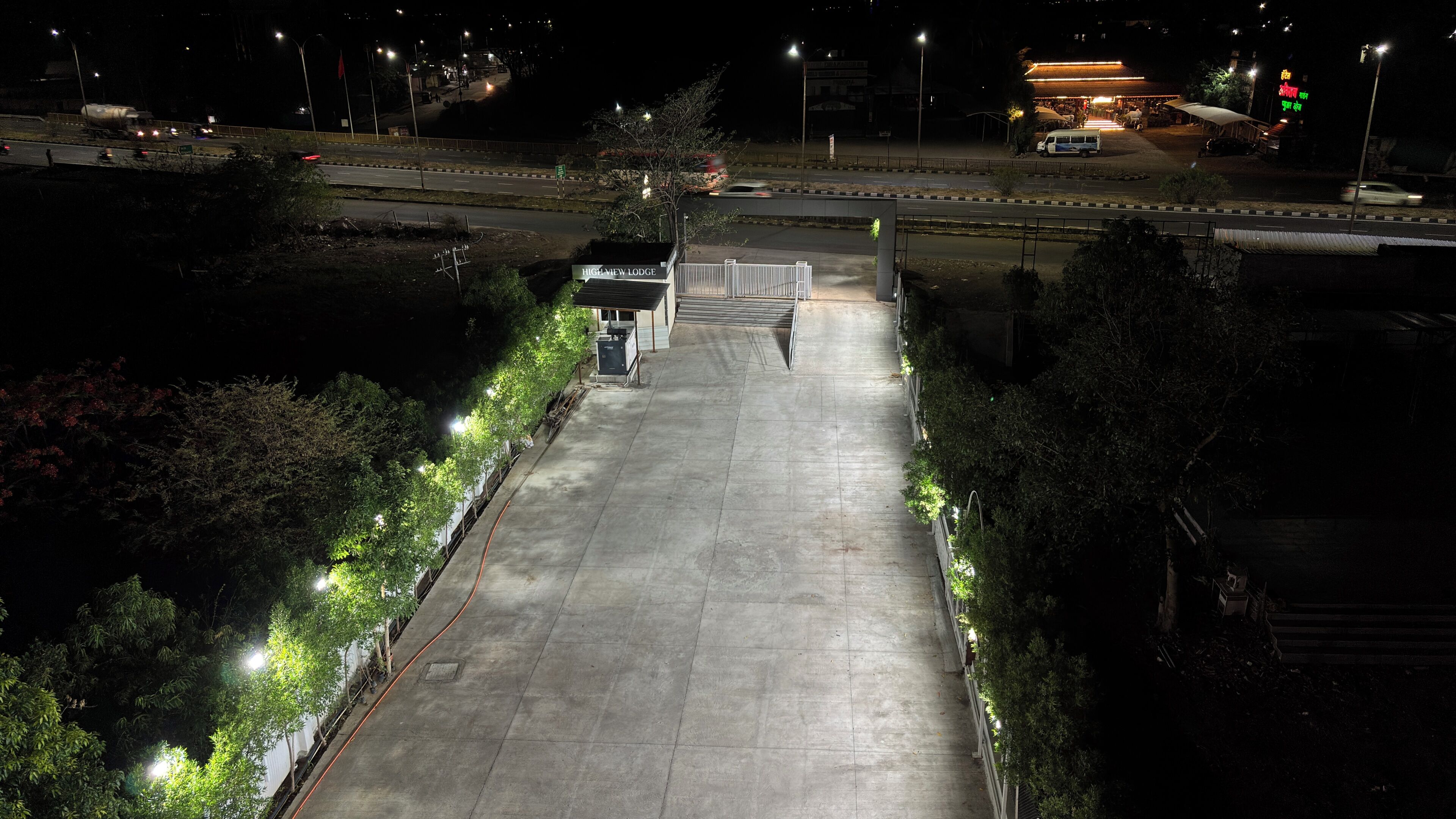 A nighttime view of a hotel entrance with a gated driveway, a sign for 'Hugh New Lodge,' and a road with vehicles.