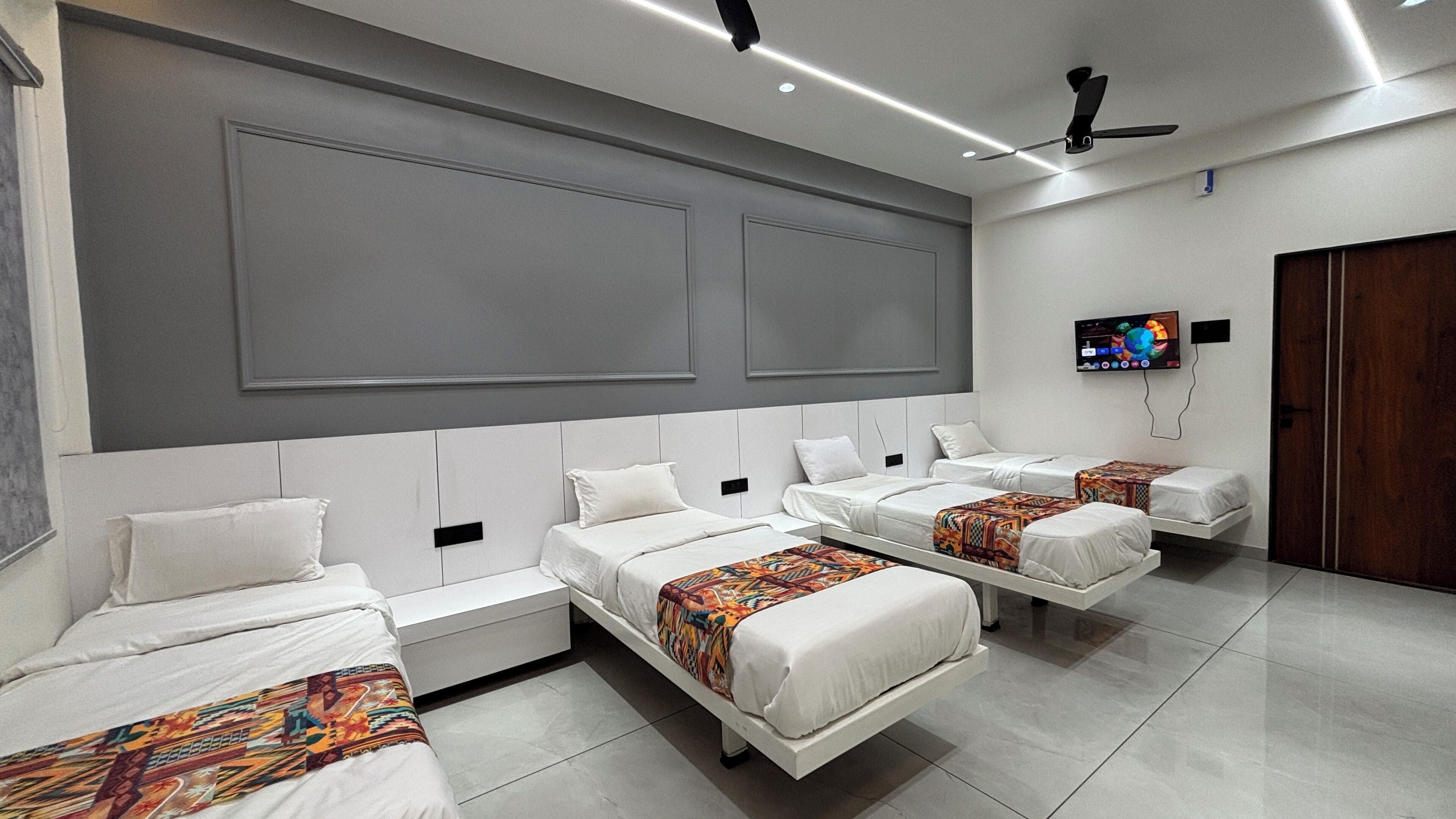 A hotel room with four beds arranged in a row, a flat-screen TV mounted on the wall, and a ceiling fan.