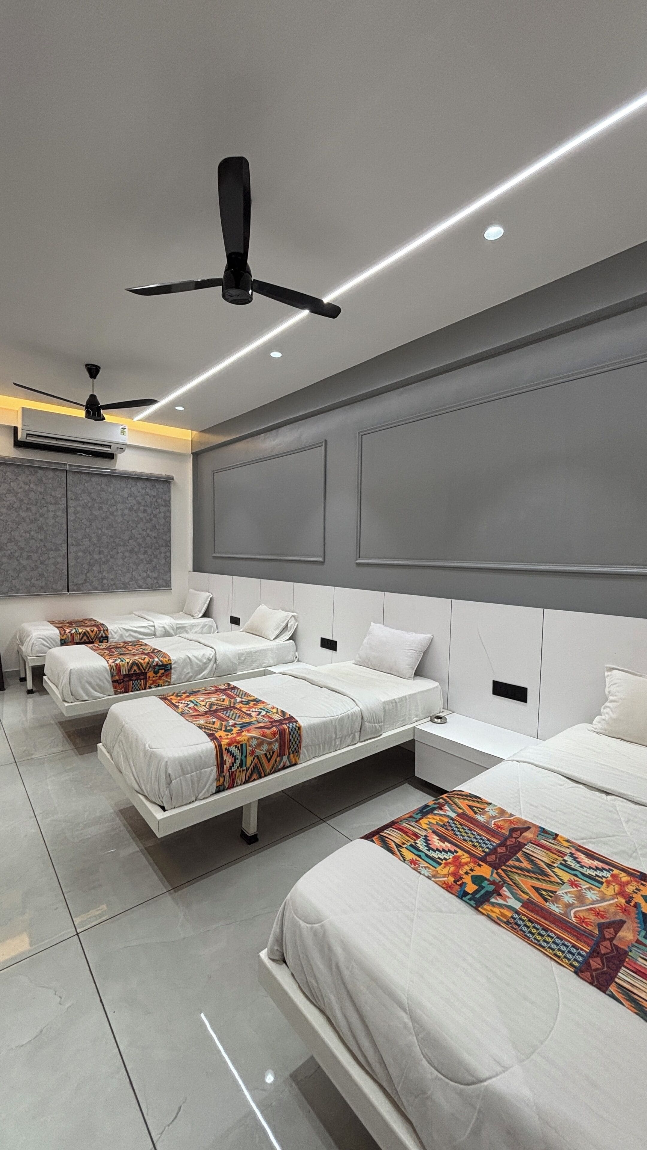 A modern hotel room with three beds, a ceiling fan, recessed lighting, and a large window with blinds.