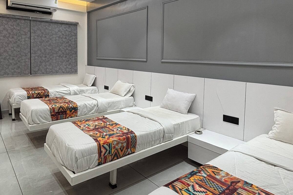 A modern hotel room with three beds, a ceiling fan, recessed lighting, and a large window with blinds.