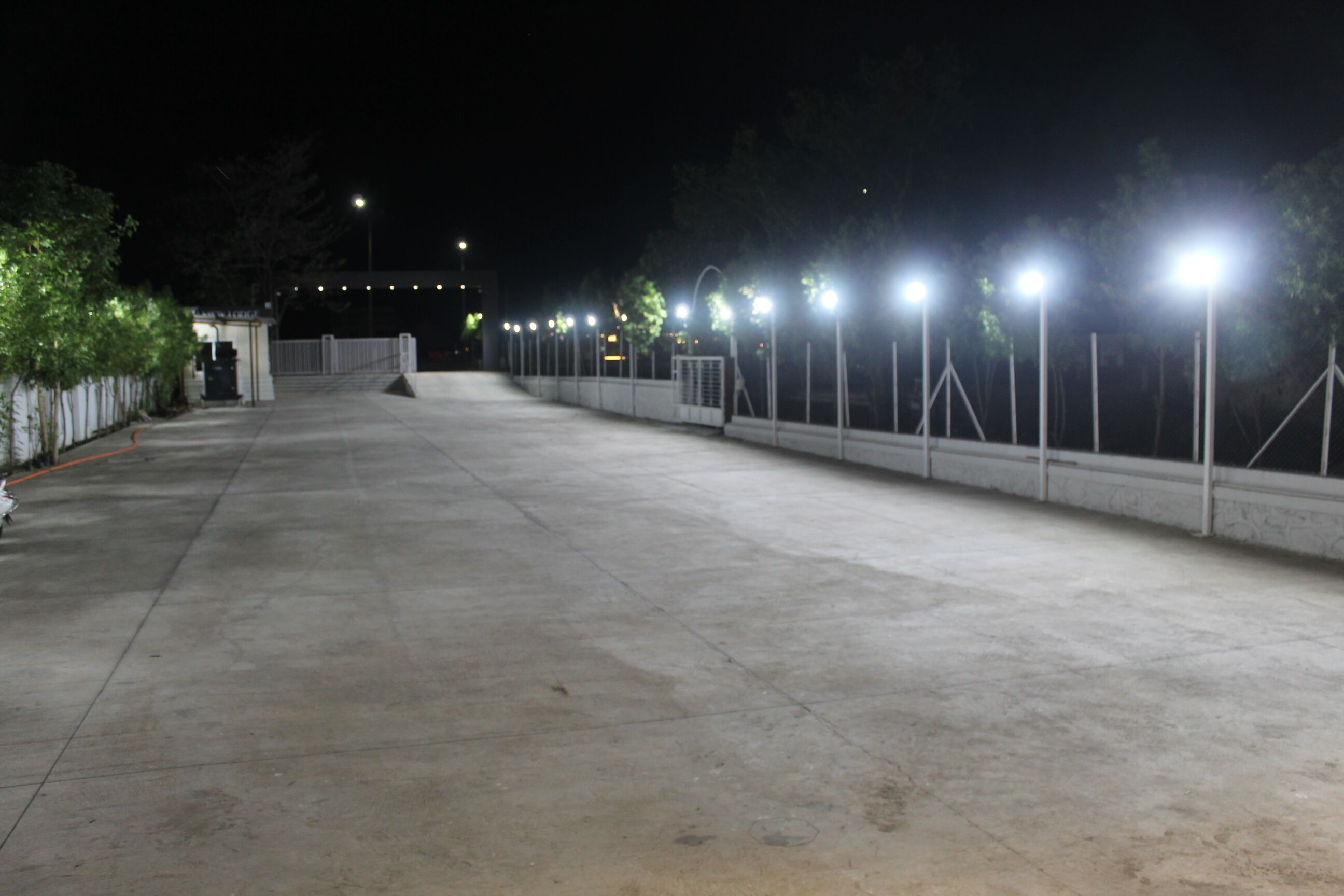 A wide, open concrete area with a series of evenly spaced lights along the perimeter, a white fence, and a small structure in the distance.