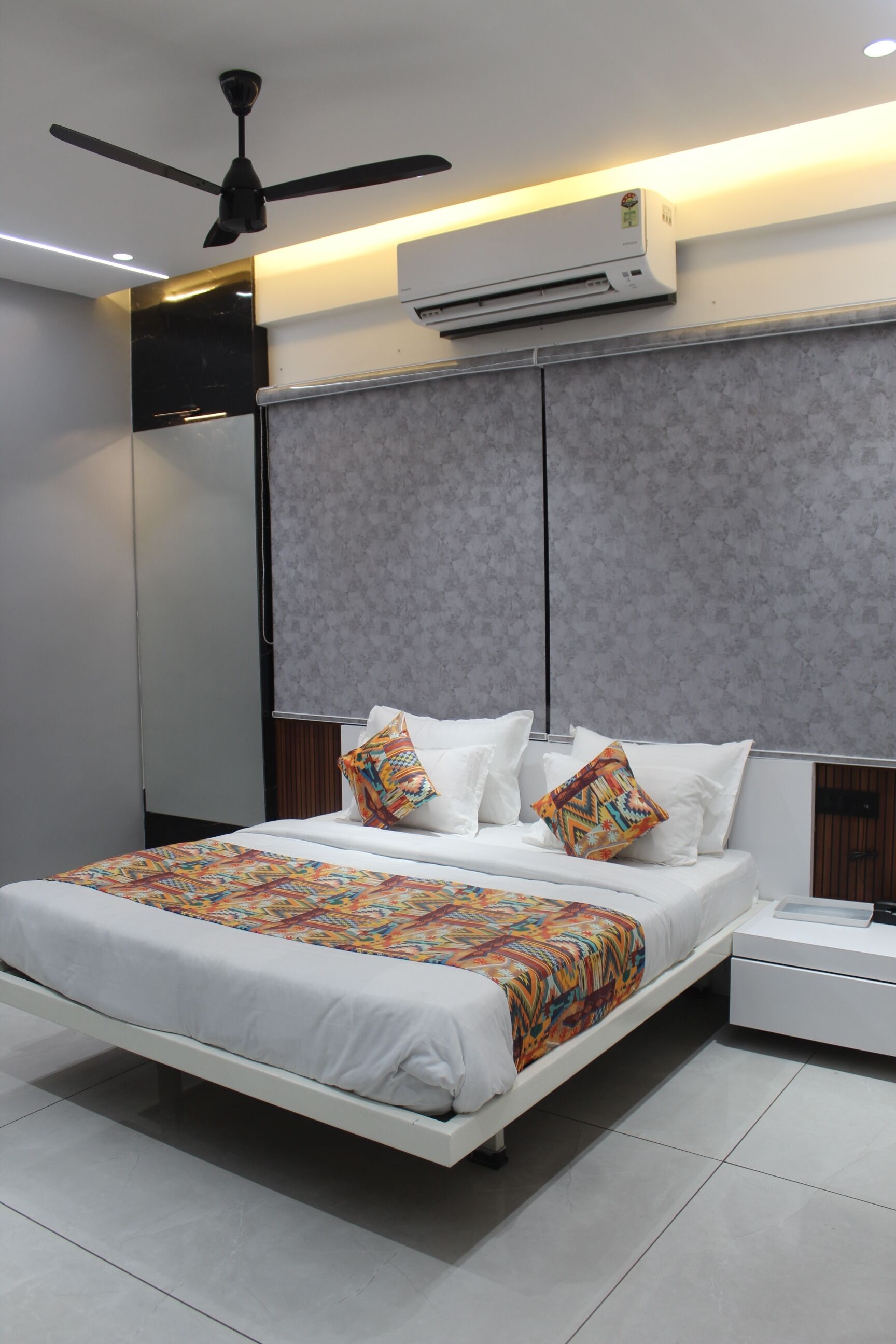 A modern hotel room with a bed, nightstand, air conditioning unit, ceiling fan, and a patterned bedspread.